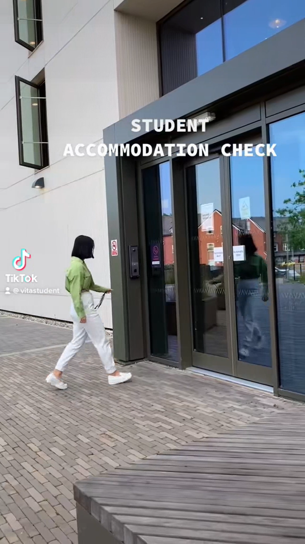 A person in white pants and a green jacket approaches the glass entrance of a building labeled "Student Accommodation Check," possibly preparing for studying abroad. The setting includes a brick walkway and reflections of buildings reminiscent of India's vibrant architecture.